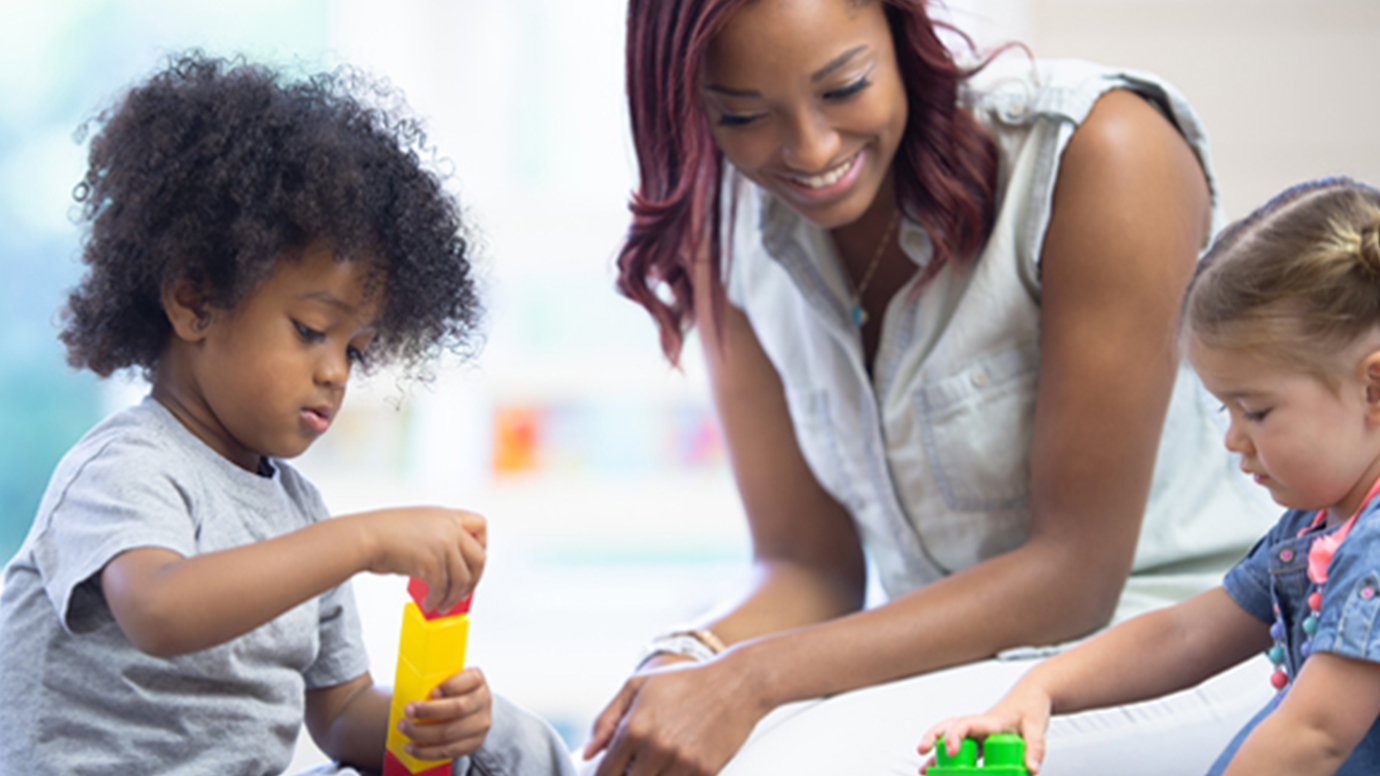 An African American teacher helps two toddlers play with blocks.